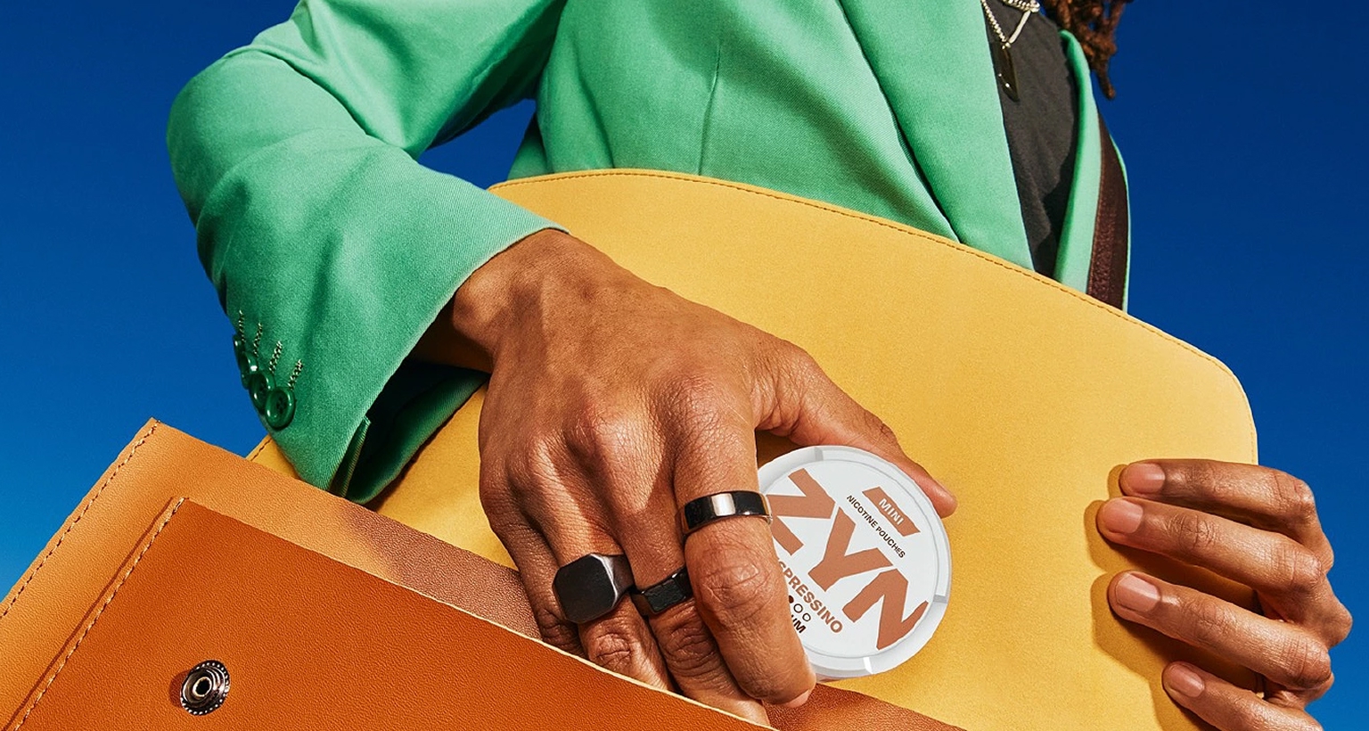 Person steckt eine ZYN-Dose in eine gelbe Ledertasche, vor klarem blauem Himmel.