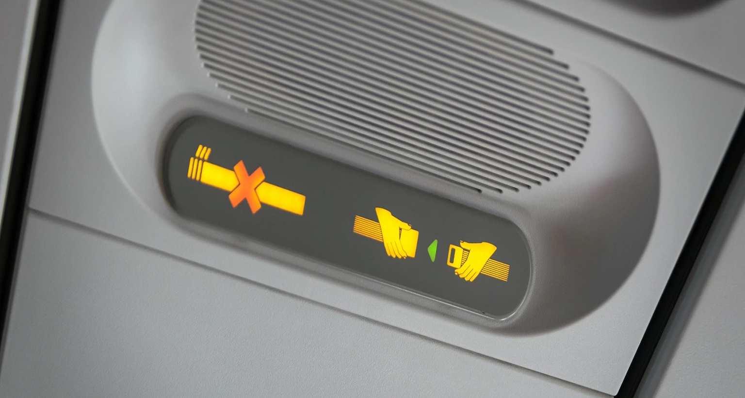 Nahaufnahme der Anzeigenleiste im Flugzeug mit leuchtenden Symbolen für „Anschnallen“ und „Rauchen verboten“.