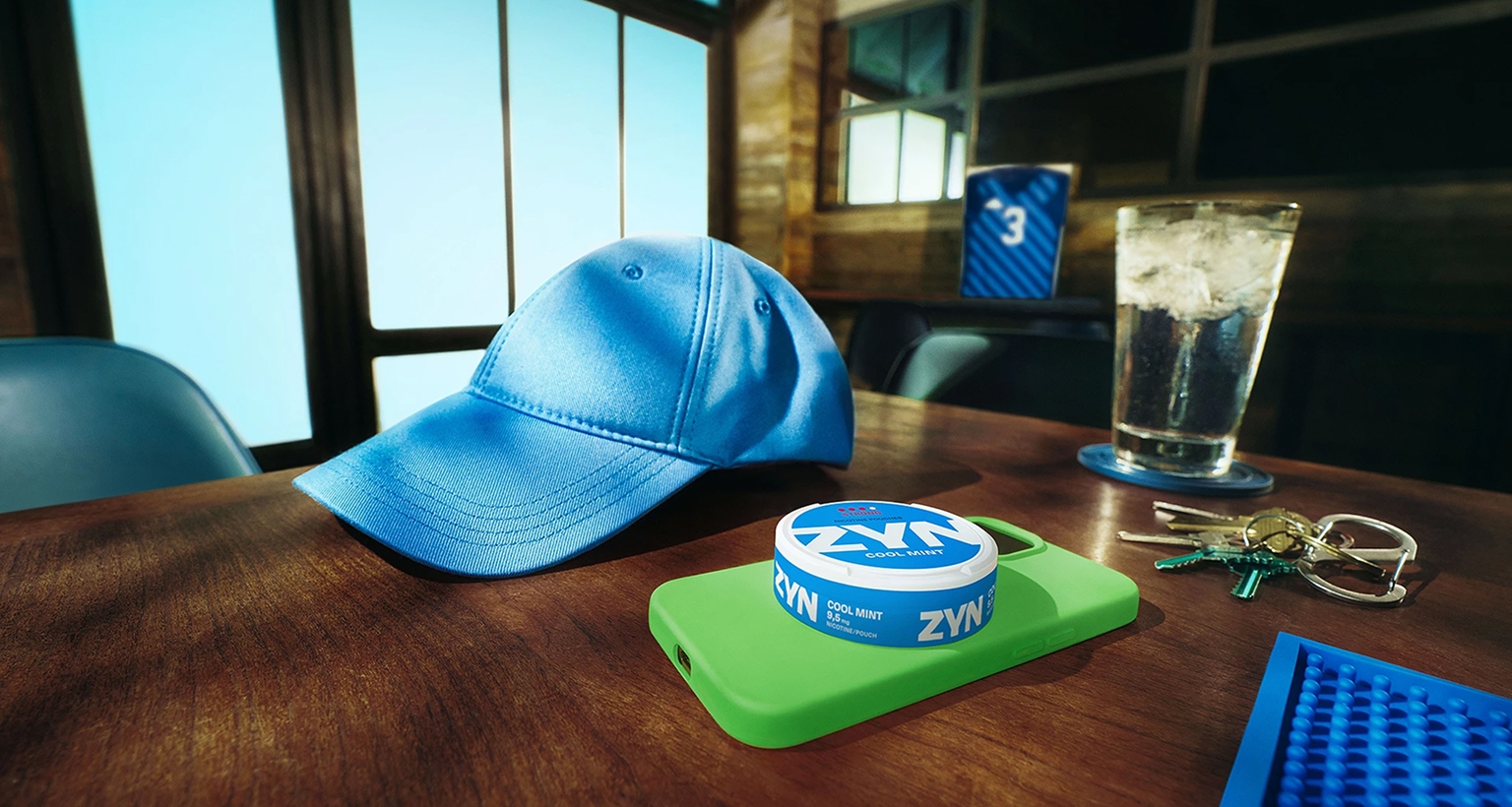 Nahaufnahme eines Holztisches mit blauer Baseballkappe, ZYN-Dose auf grünem Smartphone und Autoschlüssel im Hintergrund