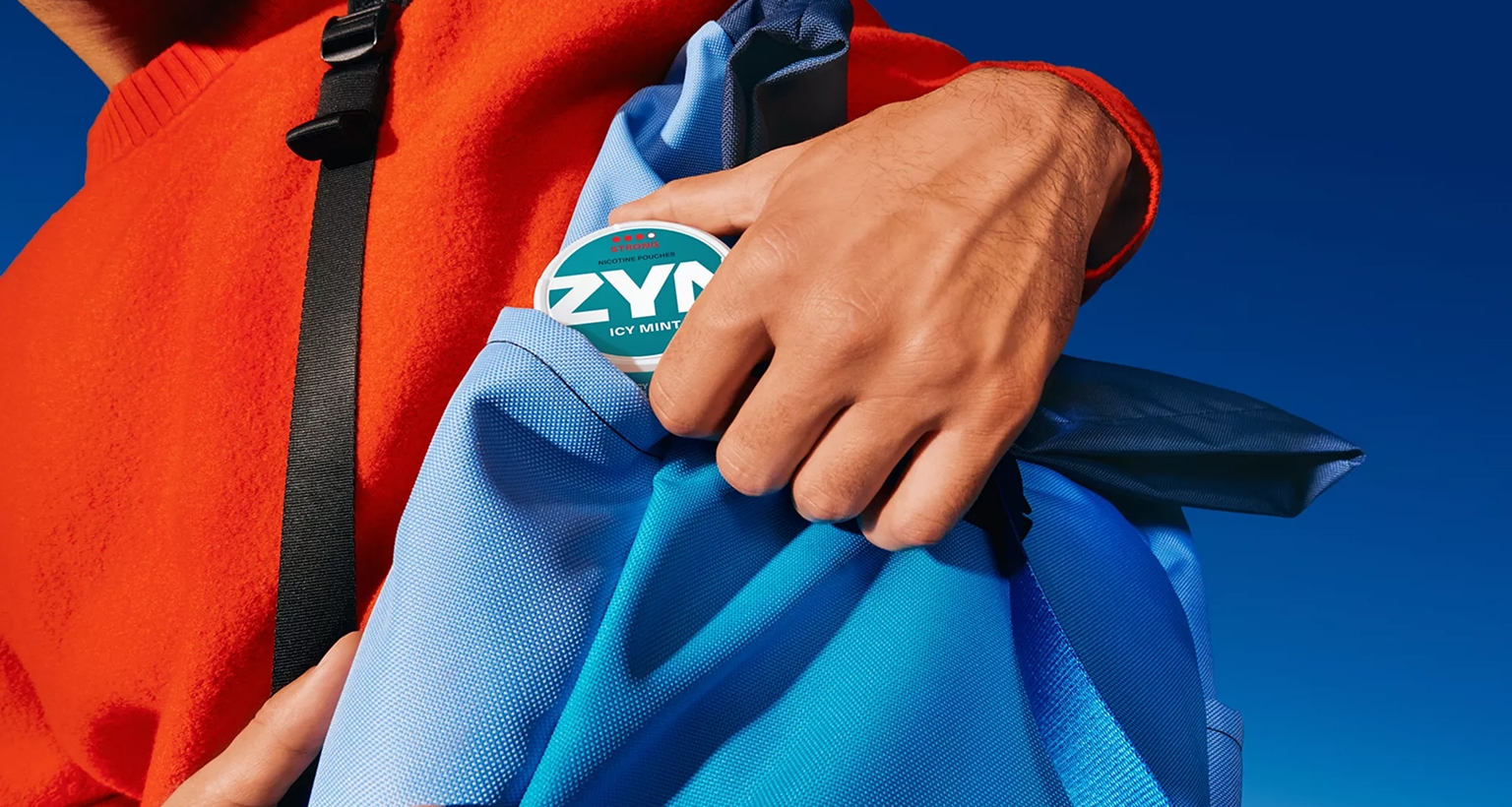 Person steckt eine ZYN-Dose in eine blaue Tasche, vor klarem blauem Himmel, trägt auffälliges rotes Oberteil.