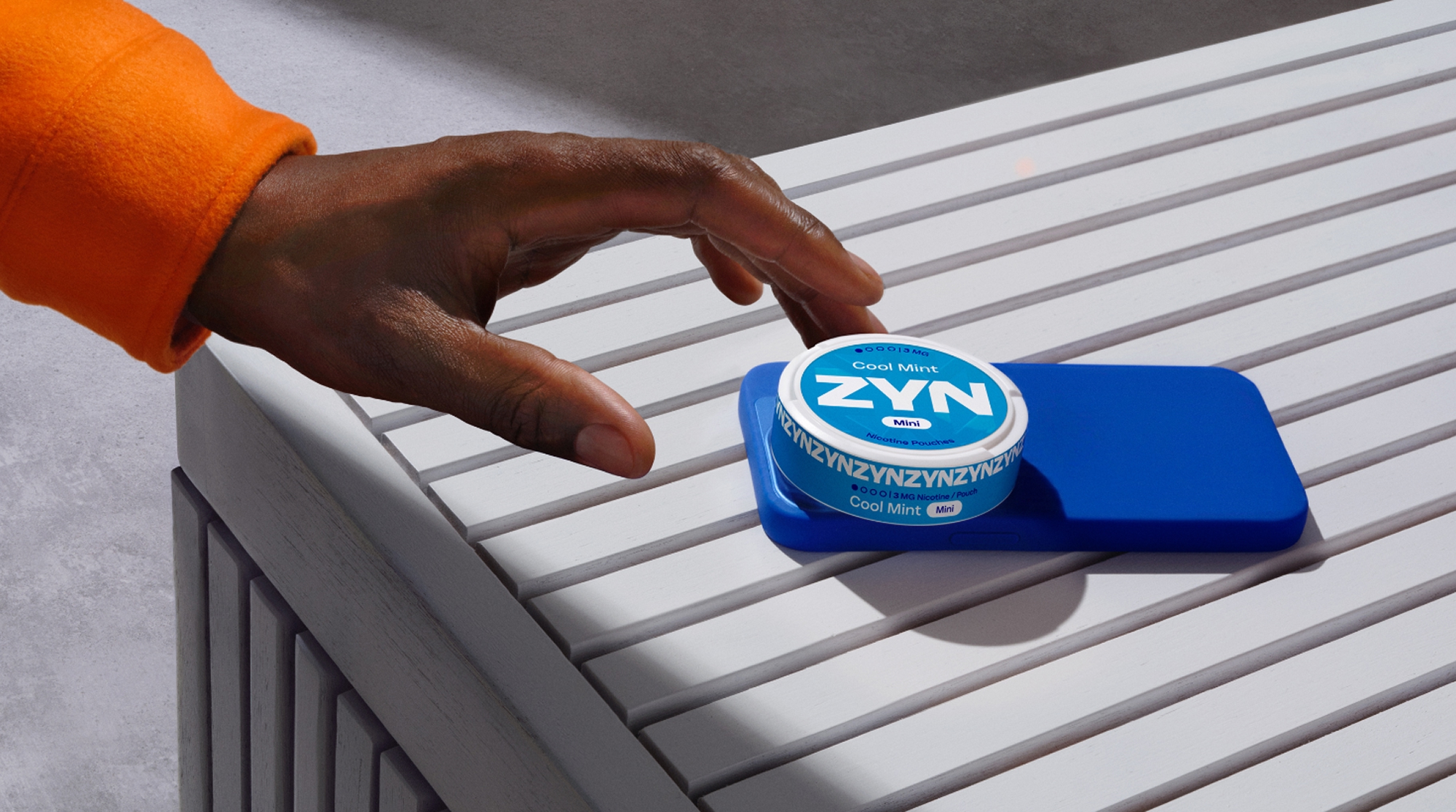 Hand greift nach einer ZYN-Dose, die auf einem Smartphone auf einer weißen Oberfläche liegt. 