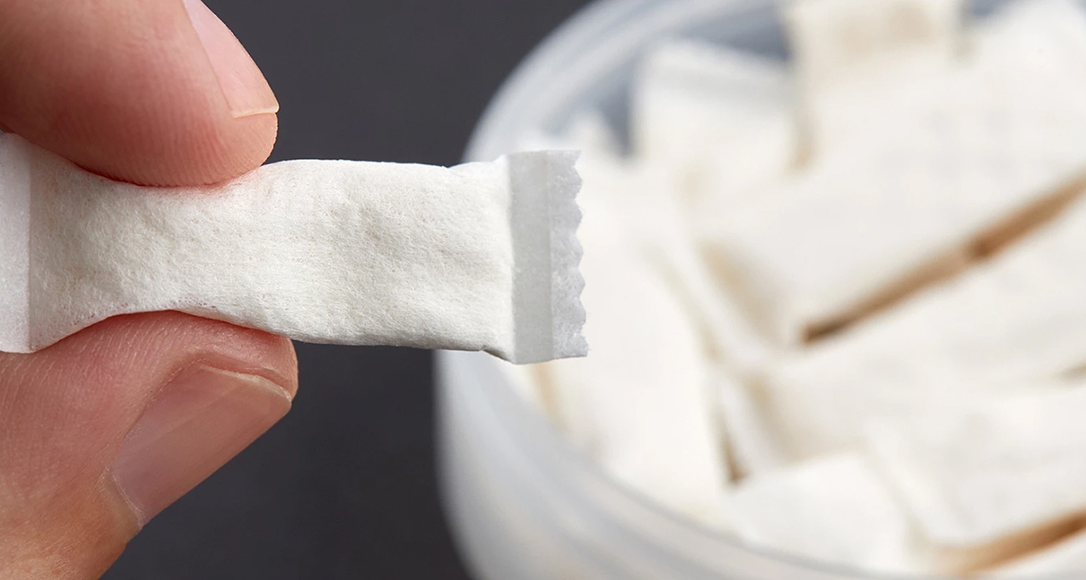 Nahaufnahme einer Hand, die ein einzelnes weißes Nicotine Pouch hält, im Hintergrund eine geöffnete Dose mit weiteren Pouches.