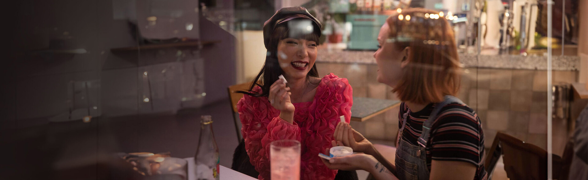 Two women sitting at bar table. They are holding ZYN nicotine pouches and a ZYN can in their hands.