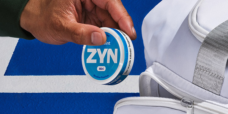 Dłoń wyjmująca opakowanie saszetek nikotynowych ZYN Cool Mint z kieszeni białego plecaka