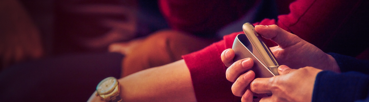 Two people sitting side by side; one holding an IQOS device.